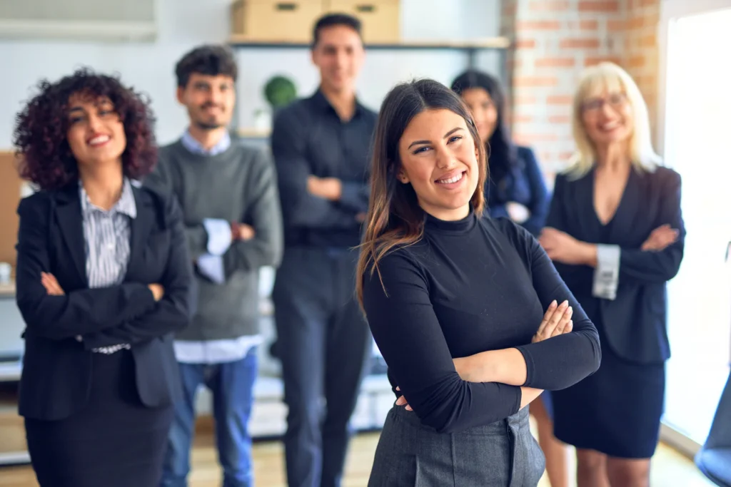 Eine Gruppe von fünf Menschen in formeller Kleidung steht zusammen in einem modernen Büro. Die Frau im Vordergrund, mit einem breiten Lächeln und den Armen verschränkt, trägt ein schwarzes Oberteil und graue Hosen. Die anderen Personen im Hintergrund, die leicht verschwommen sind, tragen ebenfalls formelle Kleidung. Das Büro hat helle Wände und eine gemütliche Atmosphäre.